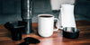 A white Ember Mug sits on a dark wooden table next to a white electric kettle and a black Aeropress with it's accessories.