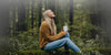 Woman in yellow sweater sitting in a forest and looking up while holding a white Ember Mug².